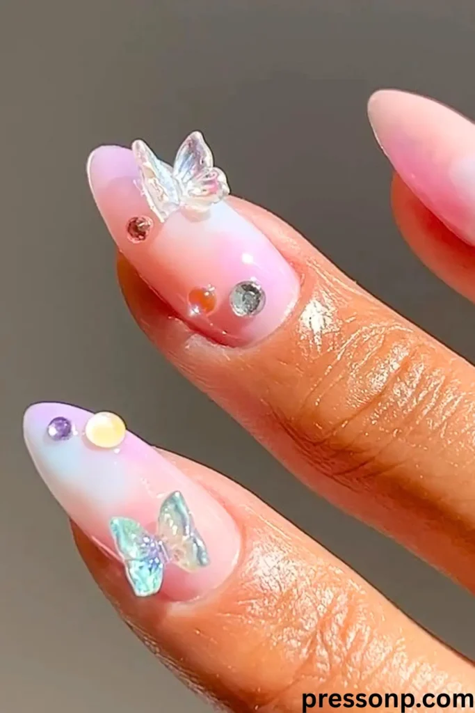 A close-up of almond-shaped nails featuring a soft, sheer pink and blue gradient base. The design is accented with small, iridescent 3D butterfly charms and colorful miniature rhinestones.