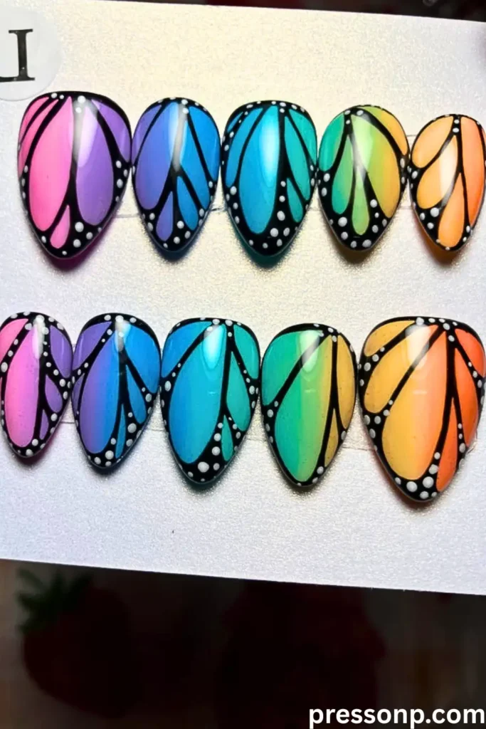 A set of press-on nails featuring a "Butterfly Wing Ombré" design with vibrant gradients in pink, blue, teal, yellow, and orange, finished with detailed black wing veins and white dots.