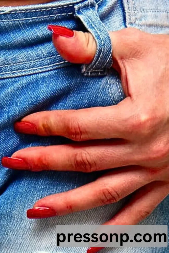 A close-up of a hand with long, vibrant red manicured nails resting against the pocket of blue denim jeans, demonstrating a bold color contrast.