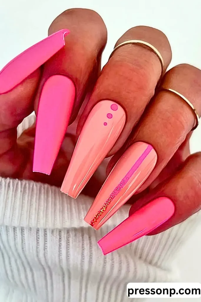 A hand with long, coffin-shaped nails in various shades of neon and bubblegum pink, featuring minimalist nail art and thin gold rings, posed against a white ribbed sweater.