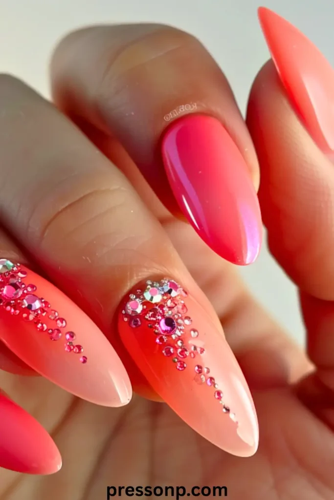 A close-up of long, almond-shaped nails featuring a "Coral Dream" manicure with a vibrant pinkish-coral gradient and detailed pink rhinestone embellishments at the cuticles.