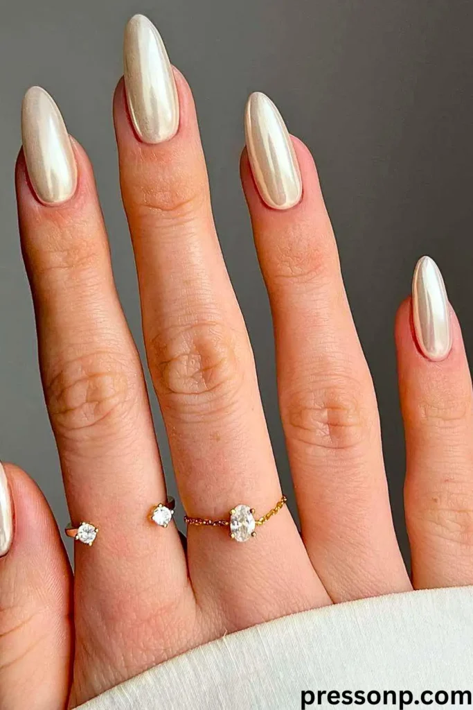A close-up of a hand with long, oval-shaped nails featuring a "Bare Beauty" glazed chrome manicure with a pearlescent white finish.