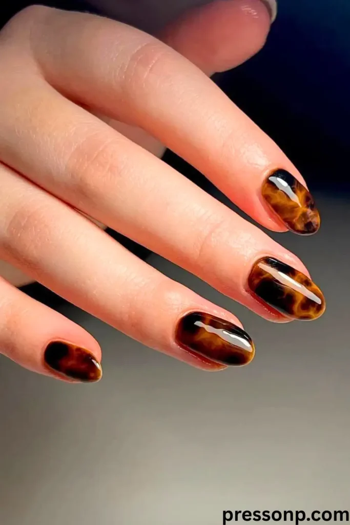 A close-up of a hand with elegant, short oval nails featuring a high-gloss, rich amber and dark brown tortoiseshell pattern.