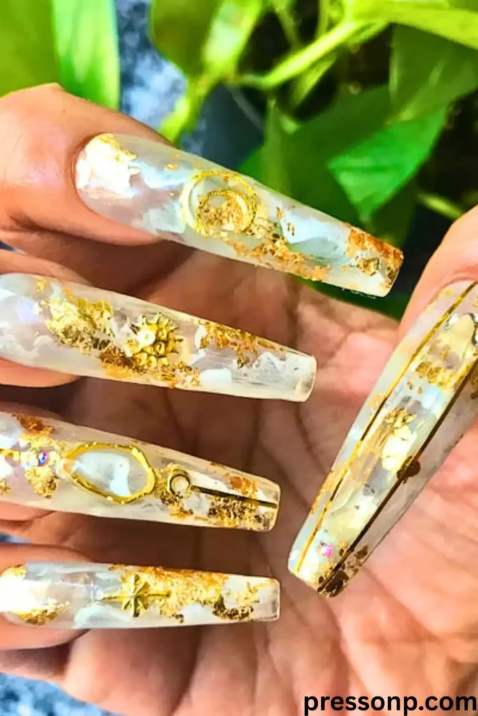 A close-up of long, square-tapered acrylic nails featuring a translucent milky white quartz base embedded with luxurious gold flakes, metallic celestial charms, and fine gold outlines.