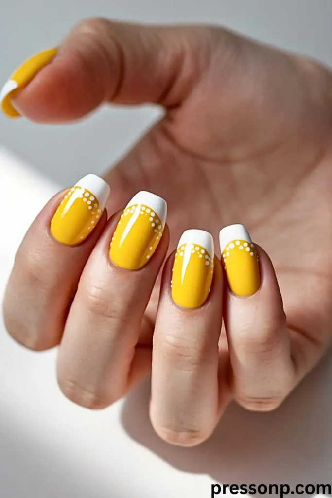 A hand featuring short, square nails with bright sunny yellow polish and a white French tip accented by delicate white polka dots.