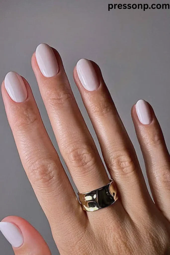 "A hand with medium skin and golden undertones featuring short, oval-shaped nails in a soft milky white polish. The creamy neutral shade provides a clean contrast that brightens warm skin tones. The hand wears a thick, hammered gold band ring, and the pressonp.com watermark is visible in the top right corner."