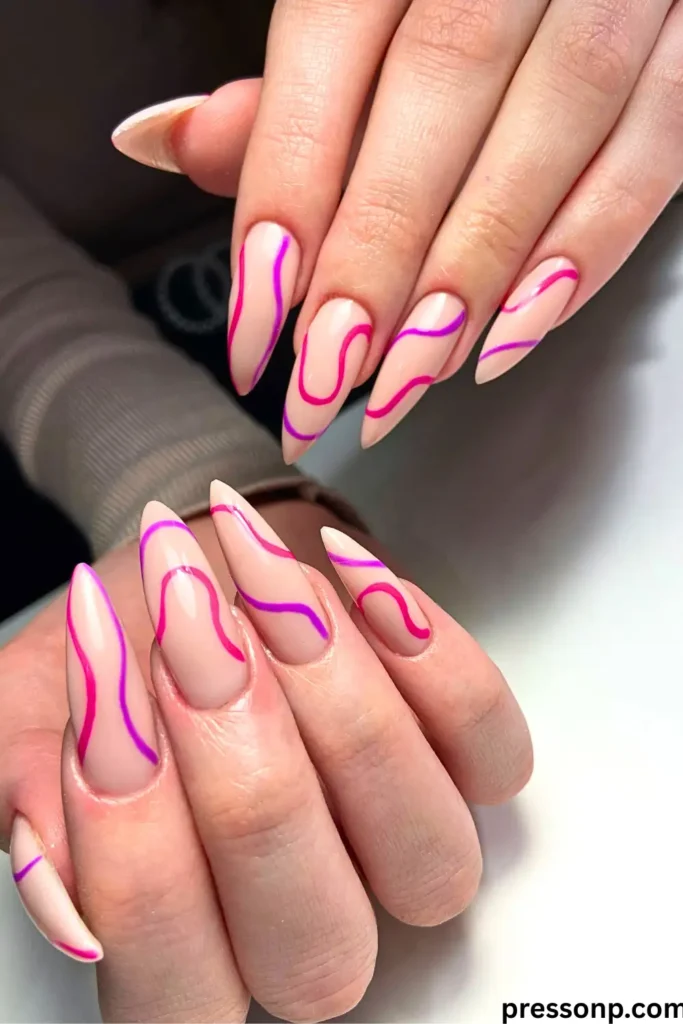 A close-up of hands displaying long, stiletto-shaped almond nails with a minimalist nude base decorated with vibrant, flowing abstract squiggles in neon coral-pink and purple.