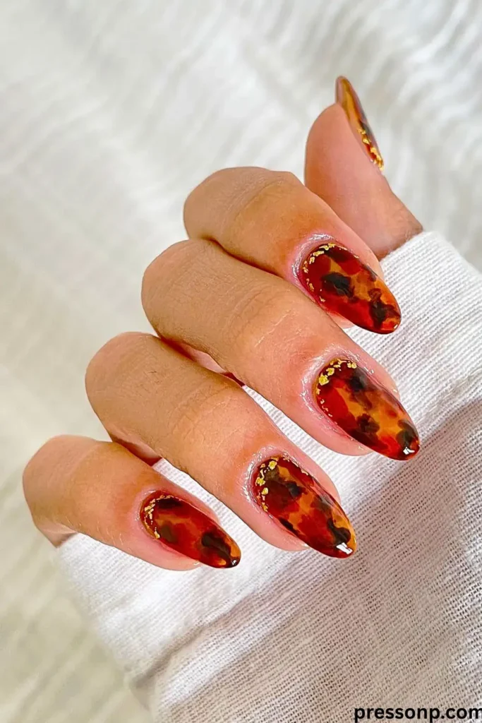 A close-up of long, square-shaped nails featuring a mixed manicure design of high-gloss amber tortoiseshell patterns and solid cream-colored accent nails decorated with gold foil flakes.
