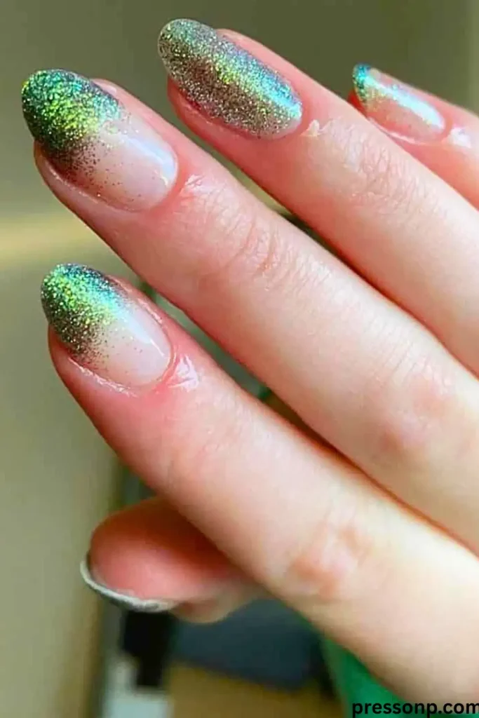 A close-up of long, oval nails featuring a "Glitter Ombré" manicure with a smooth transition from a clear, neutral nude base to dense green and silver sparkly tips.