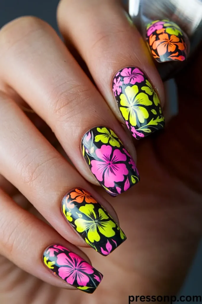 A close-up of a hand with square-shaped nails featuring a striking matte black manicure covered in vibrant, neon pink, lime green, and electric orange Hawaiian hibiscus flower patterns.