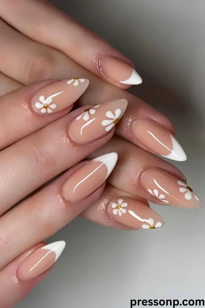 A close-up of hands with long, almond-shaped nails featuring a mixed manicure design that pairs classic crisp white French tips with delicate hand-painted white daisies over a glossy nude base.