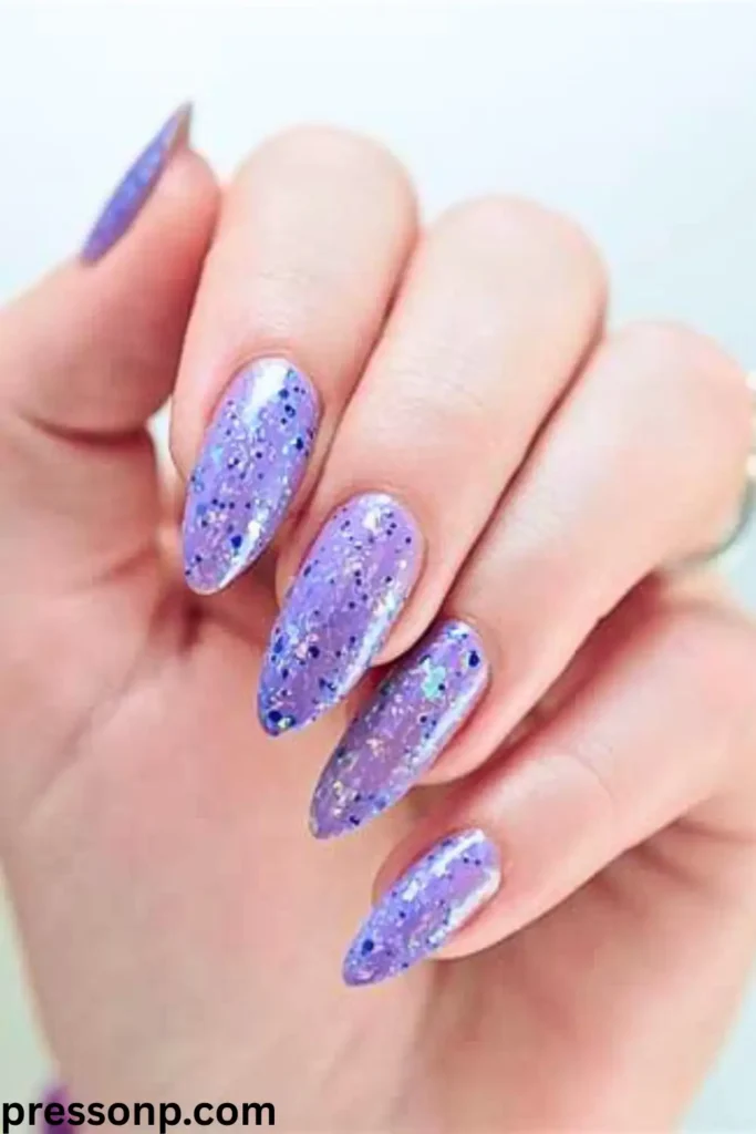 A close-up of a hand with long, almond-shaped nails featuring a "Lavender Dreams" manicure coated in a soft pastel purple polish packed with reflective silver glitter and holographic flakes.