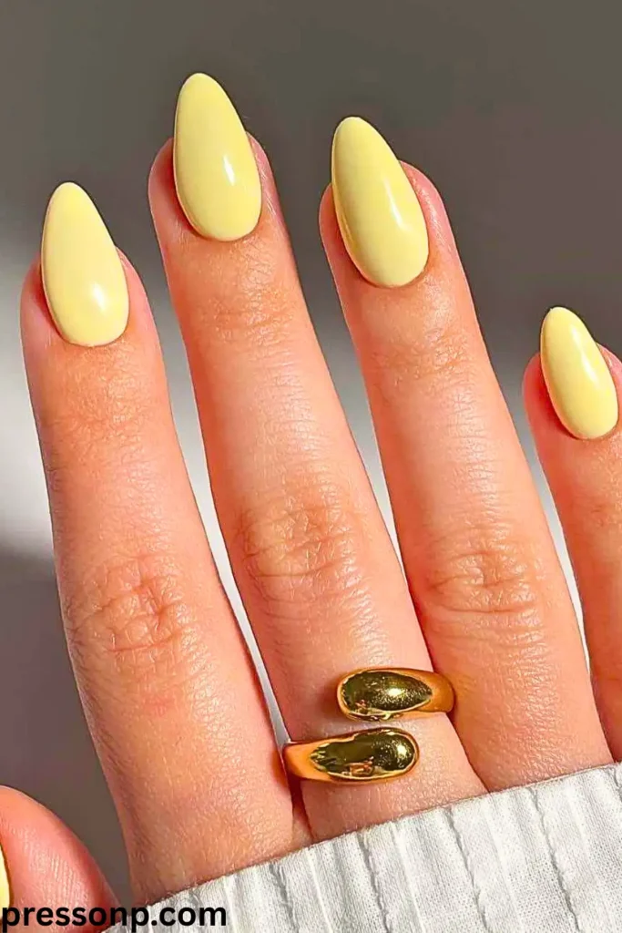 A close-up of a hand with long, almond-shaped nails painted in a smooth, high-gloss solid pastel butter yellow color, accessorized with a thick gold wrap ring.