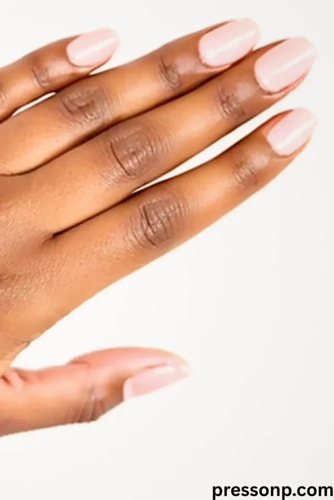 "A close-up of a hand with tan to brown skin and warm undertones featuring short, rounded square nails. The manicure uses a soft, opaque ballet pink polish that provides a bright, clean contrast against the richness of the skin tone. The pressonp.com watermark is visible in the bottom right corner."