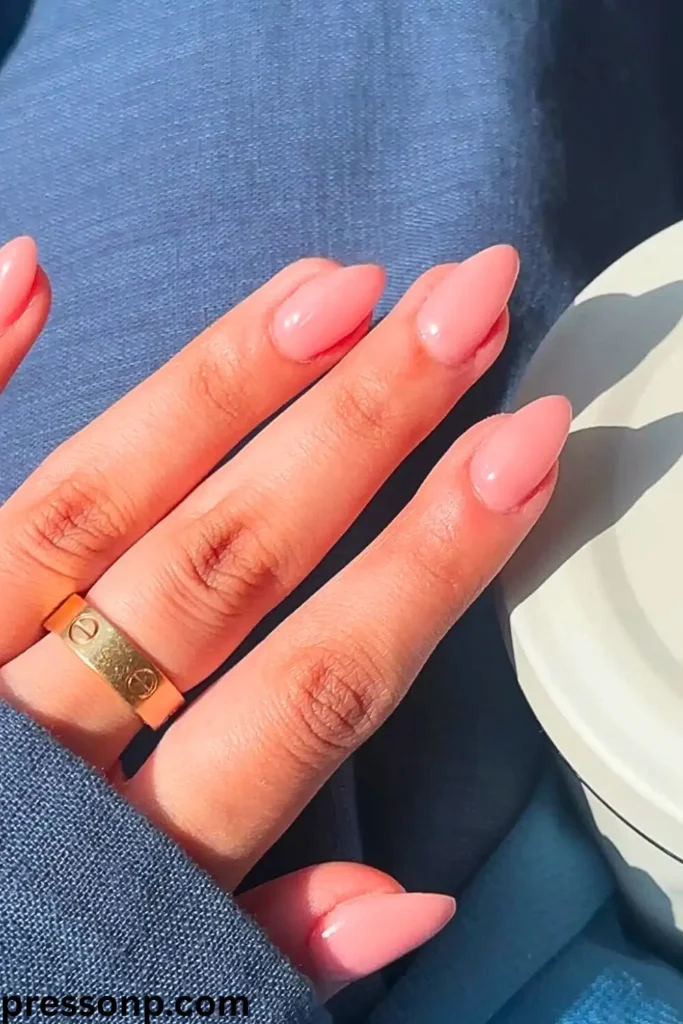 A close-up under bright sunlight of a hand with long, almond-shaped nails painted in a smooth, high-gloss solid blush pink nude polish, styled with a gold band ring.