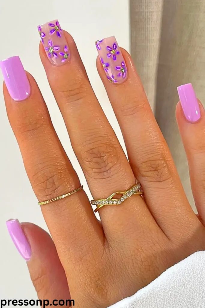 A close-up of a hand with long, square-shaped nails featuring a mix of solid pastel lilac polish and sheer accent nails decorated with hand-painted purple flowers and tiny green gems.