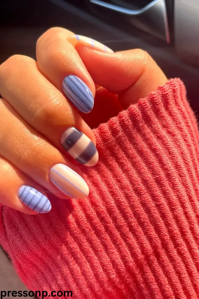 A close-up of a hand with short, round-oval nails featuring a chic striped manicure in varying shades of cornflower blue, pastel cream, and navy blue, mimicking a tailored fashion pattern.