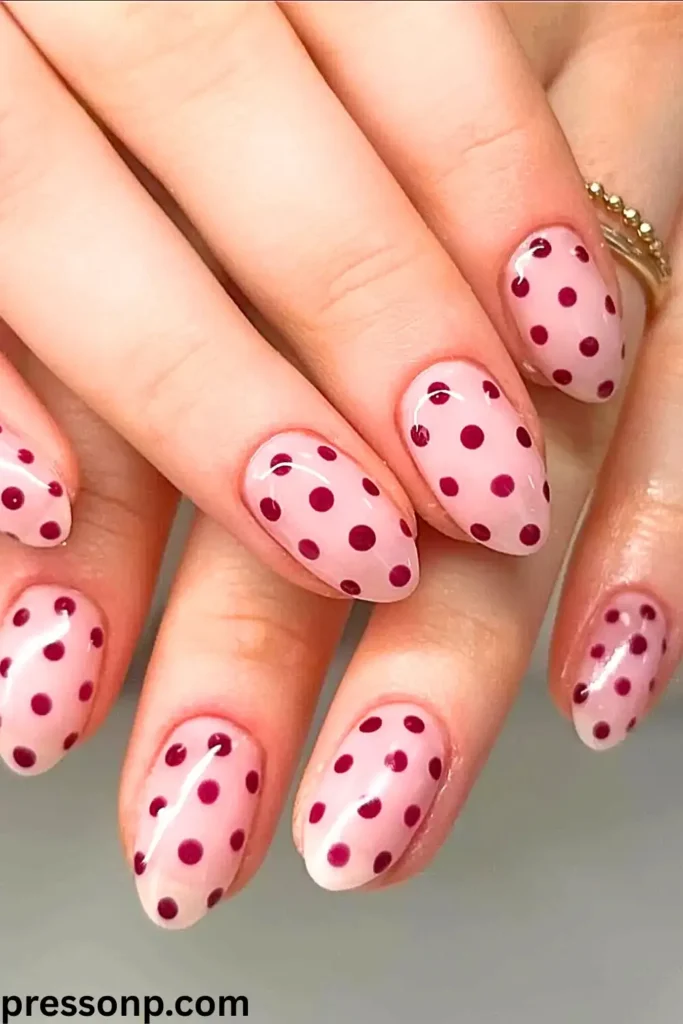 A close-up of two hands interlaced, showcasing short, rounded nails with a sweet and glossy pastel pink base decorated in a clean, uniform burgundy red polka dot pattern.