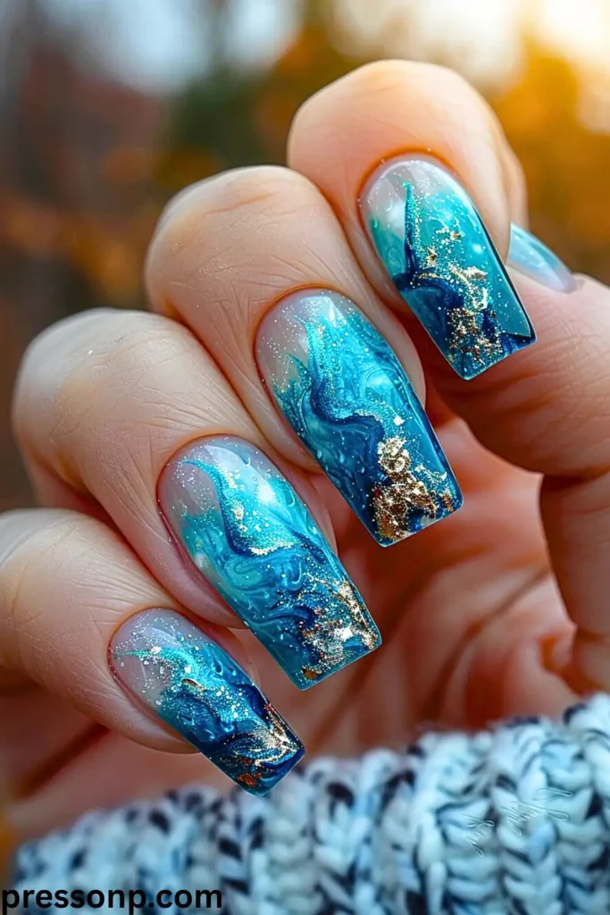 A close-up of a hand with long, square nails featuring a modern negative space ocean wave design with swirling translucent teal, deep blue, gold foil flake, and glitter accents.