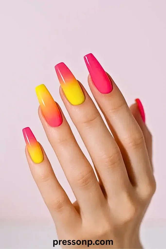 A hand with long coffin-shaped nails featuring a vibrant neon ombré gradient that blends hot pink and bright yellow, resembling a summer sunset.