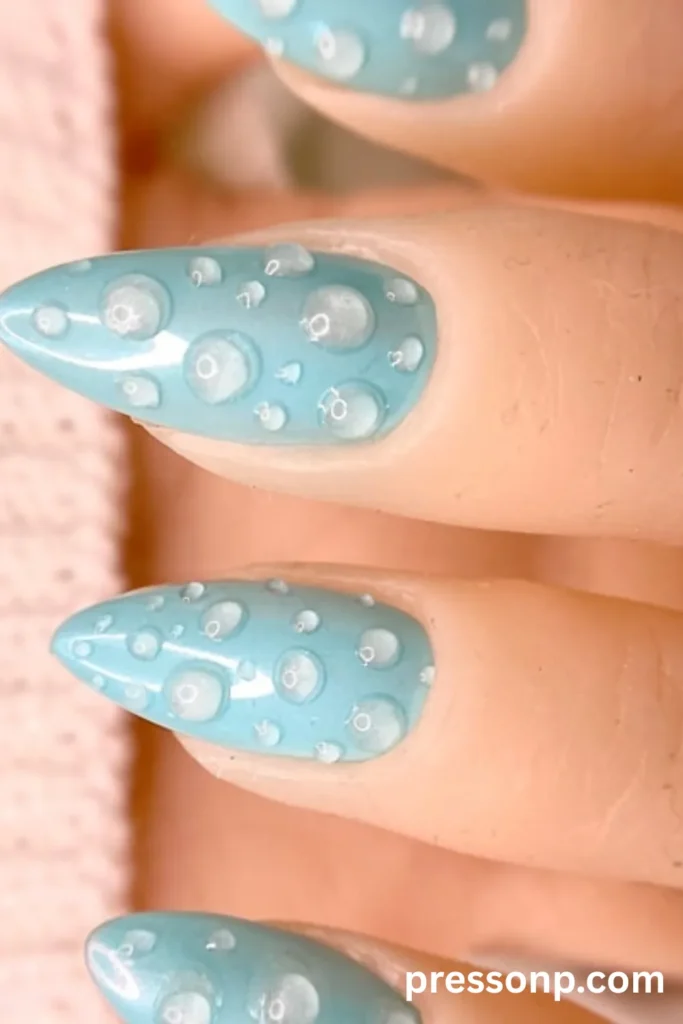Close-up of long almond-shaped nails featuring a glossy pastel blue base covered in realistic, clear 3D gel water droplets mimicking fresh dew drops.