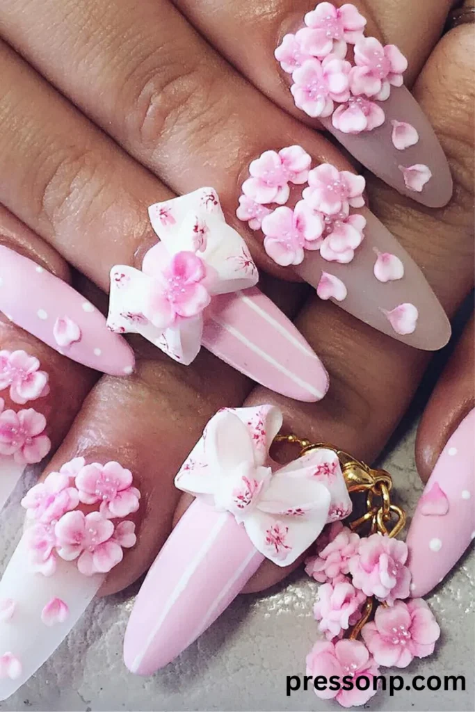 Extreme close-up of long stiletto nails featuring highly detailed pink 3D floral accents, sculpted cherry blossom petals, large structural white bows, and a hanging floral nail charm.