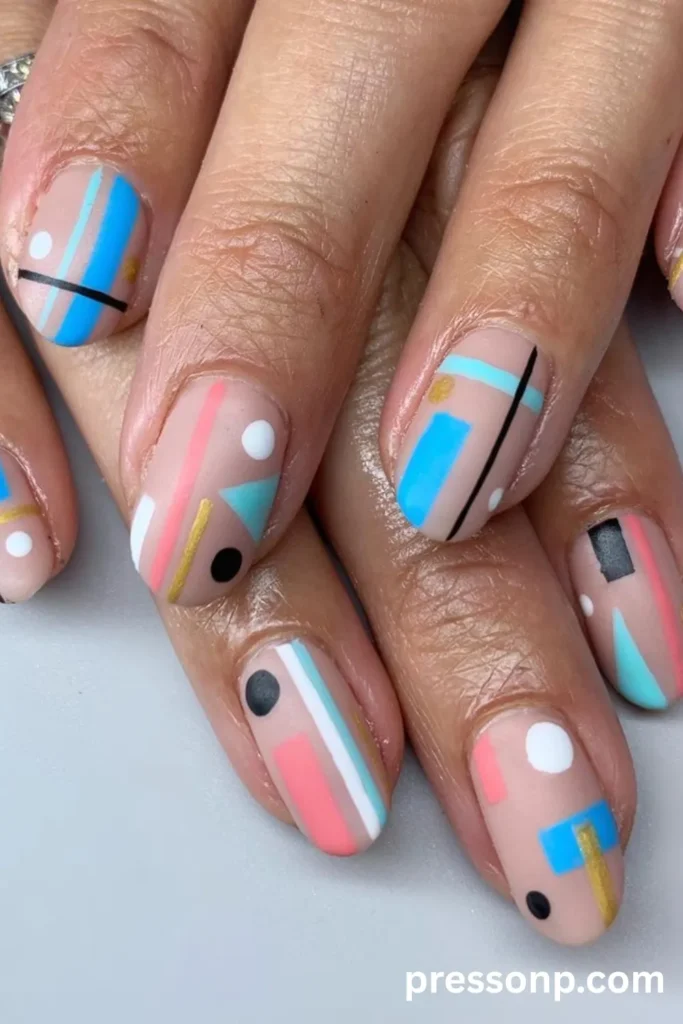Close-up of short, rounded nails with a matte nude base, featuring an abstract geometric manicure with colorful blocks, sharp lines, and dots in coral pink, bright blue, teal, and gold.