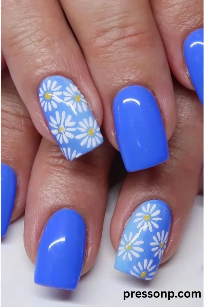 Close-up of glossy square nails featuring a solid vibrant medium blue polish paired with light blue accent nails decorated with hand-painted white daisy patterns.