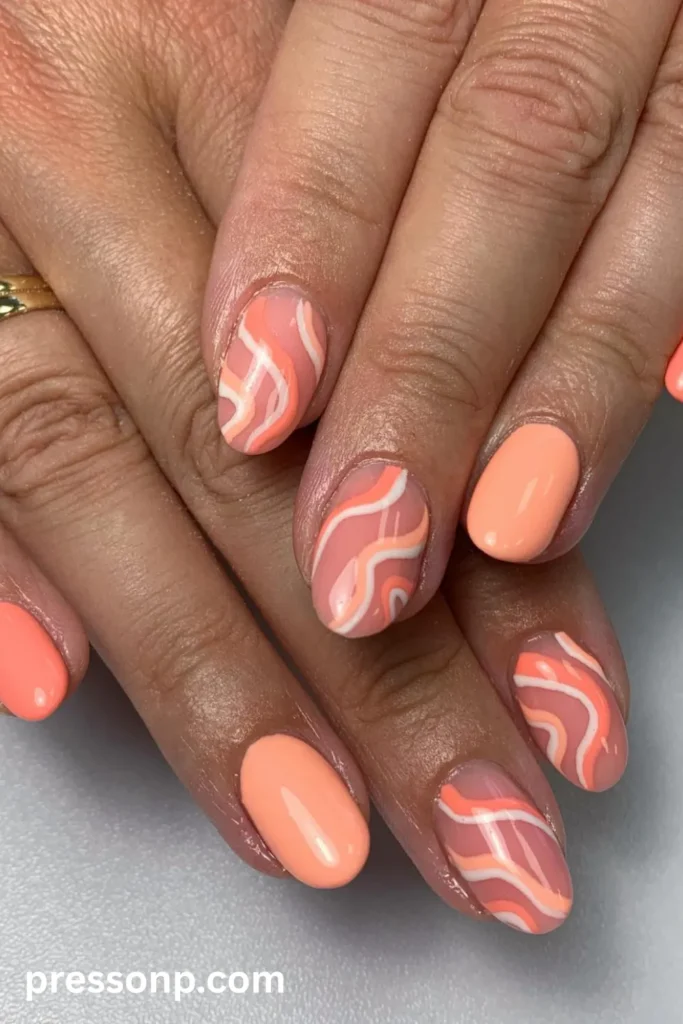Close-up of short oval nails painted in a glossy light pastel coral polish, featuring sheer accent nails decorated with retro wavy abstract swirls in bright coral, peach, and white.