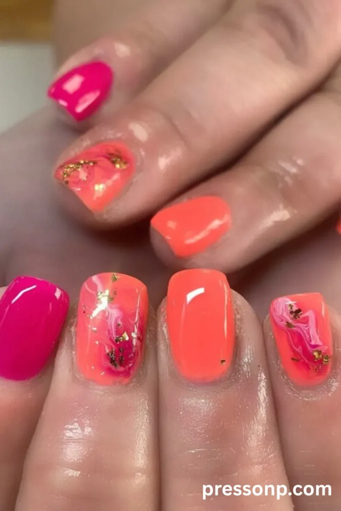 Close-up of short, square-round nails featuring a high-gloss bright coral base, mixed with hot pink accent nails and a colorful marble swirl design accented by shimmering gold leaf flecks.