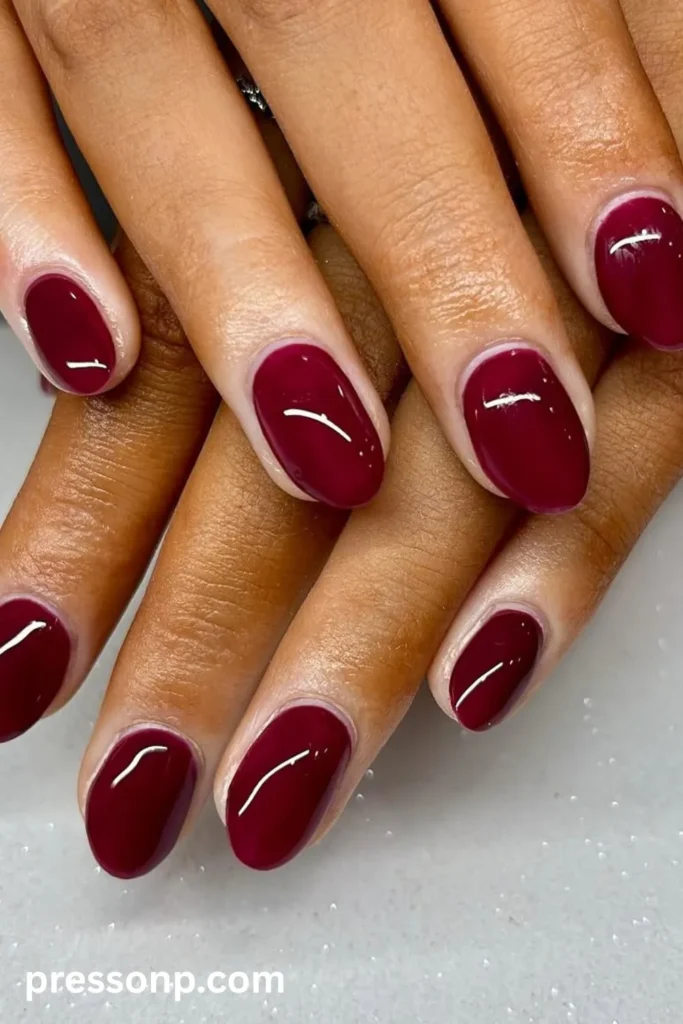 Close-up of short, neatly shaped oval nails painted in a solid, deeply saturated burgundy red polish with an ultra-reflective, glass-like glossy finish.