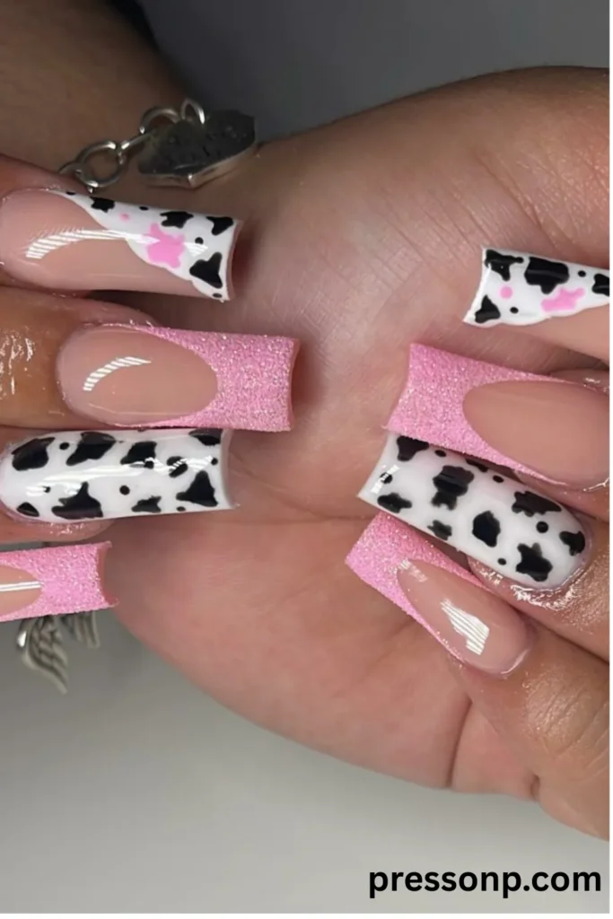 Close-up of long square nails featuring a mix of black and white cow print patterns, pink-accented cow print tips, and textured glittery pink French tips.