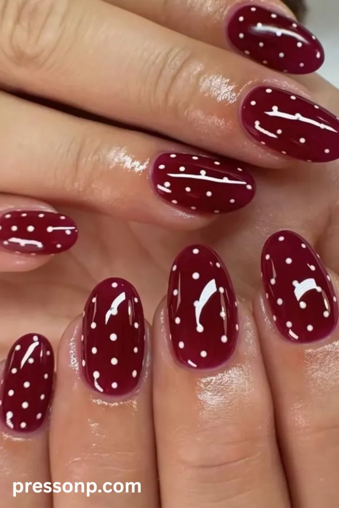 Close-up of short oval nails coated in a glossy, deep berry-burgundy polish and decorated with a uniform pattern of tiny white polka dots.