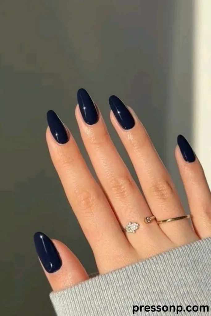 A close-up of a hand displaying long, elegant oval-shaped nails coated in a uniform, ultra-glossy deep navy blue lacquer with a glass-like shine, accessorized with delicate gold rings.