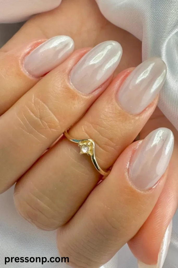 A close-up of a hand displaying an elegant glazed donut manicure on medium-length oval nails, featuring a semi-translucent milky white base topped with a pearlescent, shimmering chrome finish that gives off a tropical, seashell-like glow.