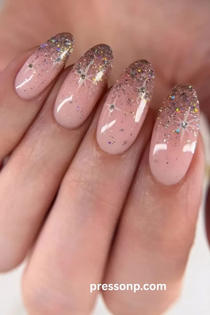 A close-up photograph of a hand displaying a soft pink manicure on long oval nails, featuring a sparkling silver and holographic glitter gradient concentrated at the tips, accented with delicate hand-painted white starbursts and tiny rhinestone centers.