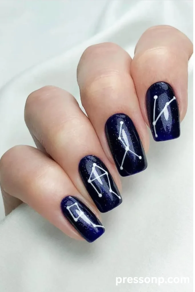Close-up of manicured nails featuring a dark blue, starry night sky base painted with crisp white constellation patterns.