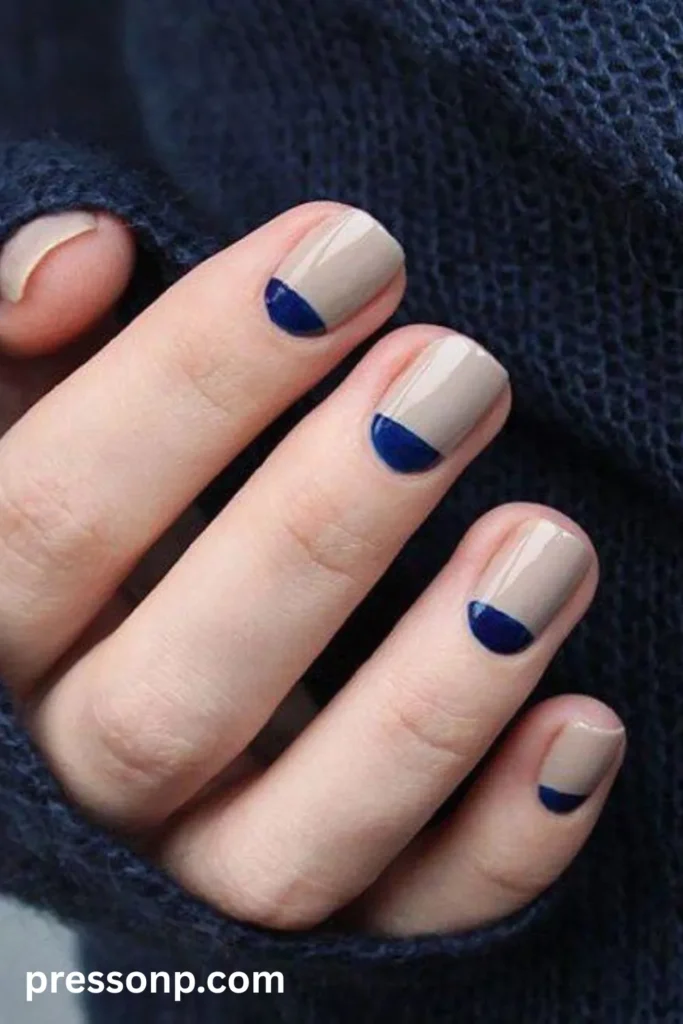 Close-up of short squoval nails featuring a glossy taupe-beige base with a stark navy blue half-moon crescent painted at the cuticle line.