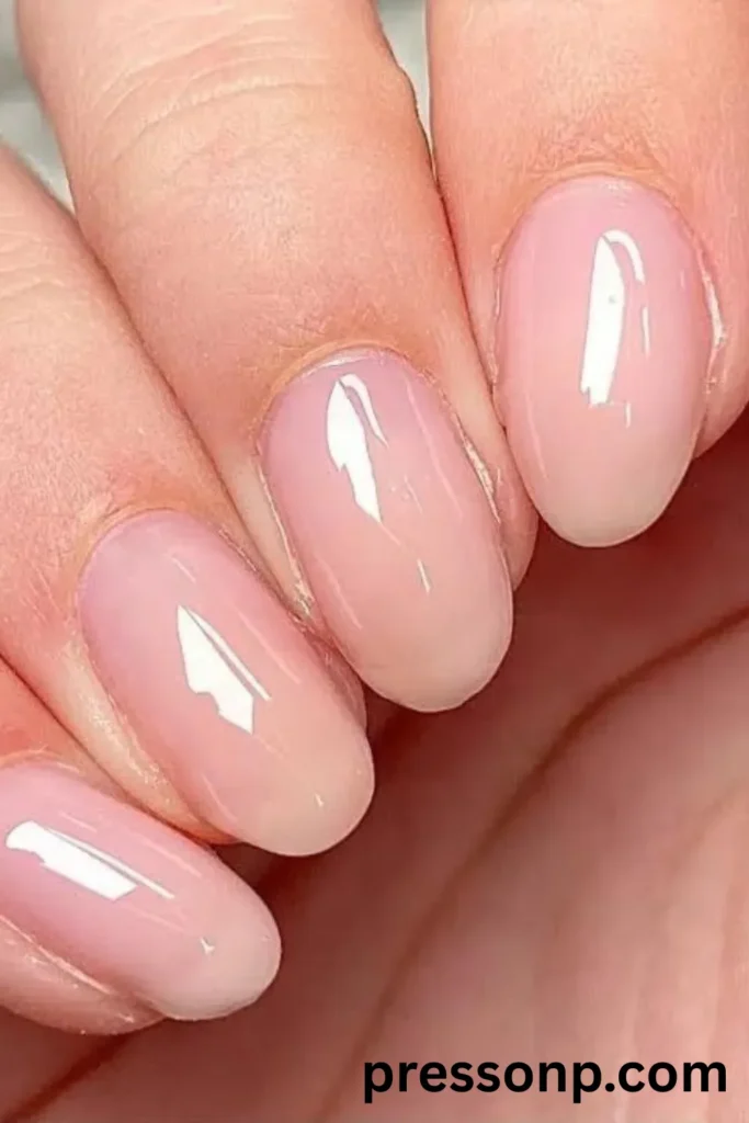 A close-up of a hand displaying short, natural oval-shaped nails coated in a solid, sheer milky-pink gel builder polish with a high-gloss, glass-like finish.