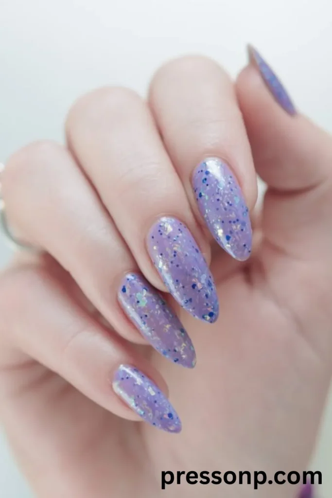 A close-up of a hand displaying long, elegant almond-shaped extensions coated in a soft pastel lavender gel lacquer, heavily layered with dimensional, iridescent glitter flakes and blue micro-specks under a high-gloss finish.