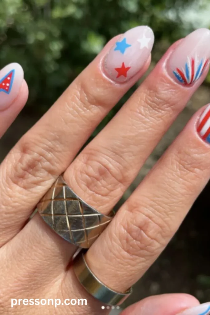 A close-up of a hand showcasing a festive red, white, and blue patriotic manicure on medium oval nails, featuring hand-painted stars, stripes, a pennant flag, and a firework burst design over a sheer milky base.