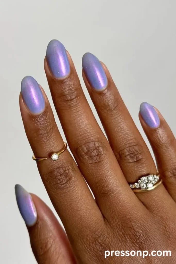 Close-up of long oval nails on a deep skin tone, featuring a luminous lavender chrome polish infused with a subtle silver shimmer and an iridescent pink hue reflection.
