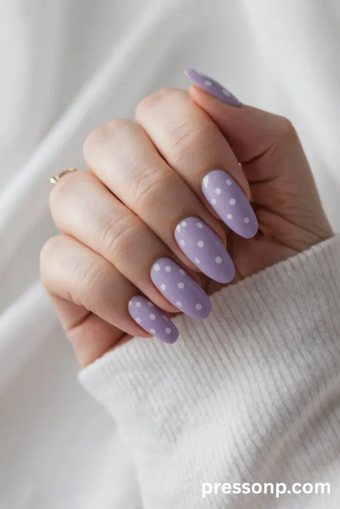 Close-up of long oval nails featuring a glossy pastel lavender base neatly decorated with a minimalist white polka-dot pattern.