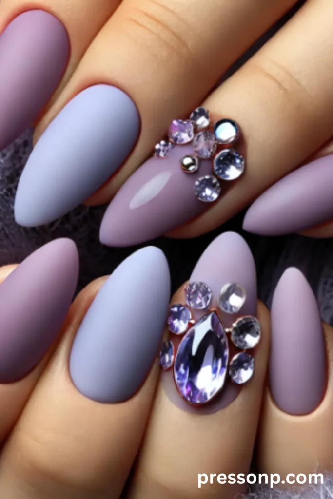 Close-up of long almond-shaped nails featuring a velvet matte finish in mixed shades of lavender and periwinkle purple, decorated with large, sparkling purple crystal clusters.