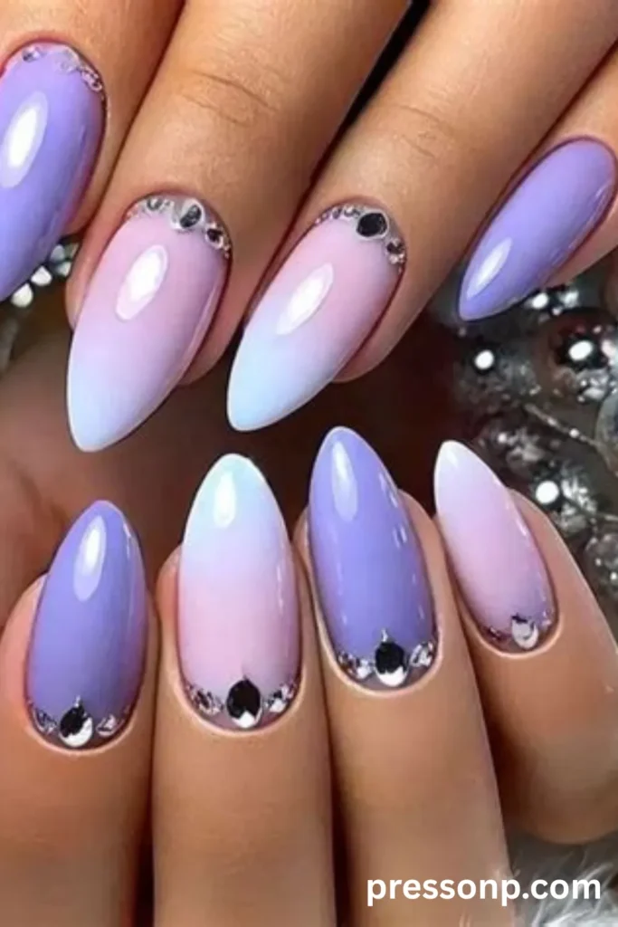 Close-up of long almond-shaped nails featuring a glossy mix of solid lavender and pastel pink-to-blue ombré gradients, accented with sparkling rhinestone clusters at the cuticles.
