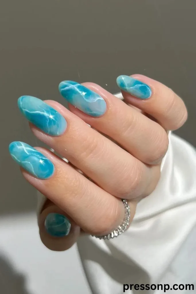 A close-up of a hand displaying long, oval-shaped extension nails with a high-gloss, water-marbled manicure featuring fluid swirls of seafoam green, turquoise, and translucent blue, accented with wispy white ripples that mimic sunlight reflecting on water.
