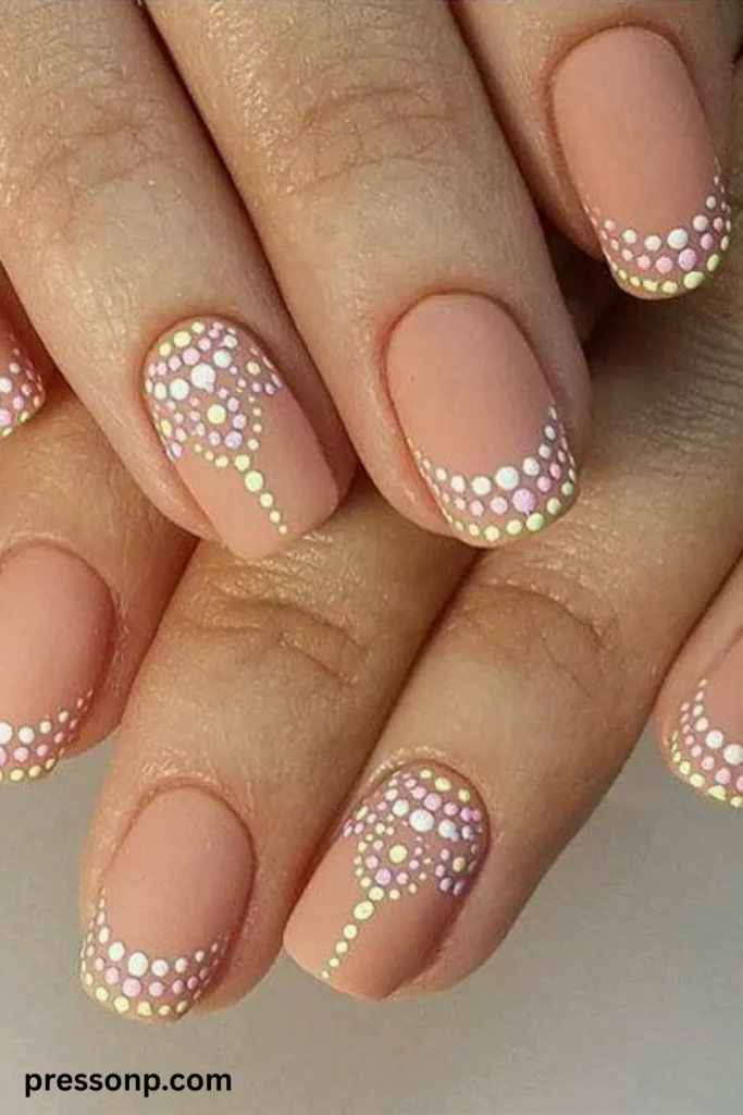 A close-up of a hand displaying a matte nude manicure on short, natural squoval nails, featuring intricate pointillism nail art made of tiny white, pink, and pale yellow dots arranged into curved French tips and delicate mandala-style patterns.