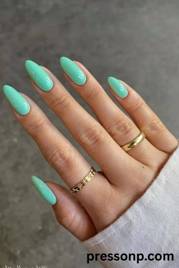 A close-up of a hand showcasing long, elegant oval-shaped extensions coated in a vibrant, solid mint green gel polish with an ultra-glossy finish, accessorized with two gold bands.