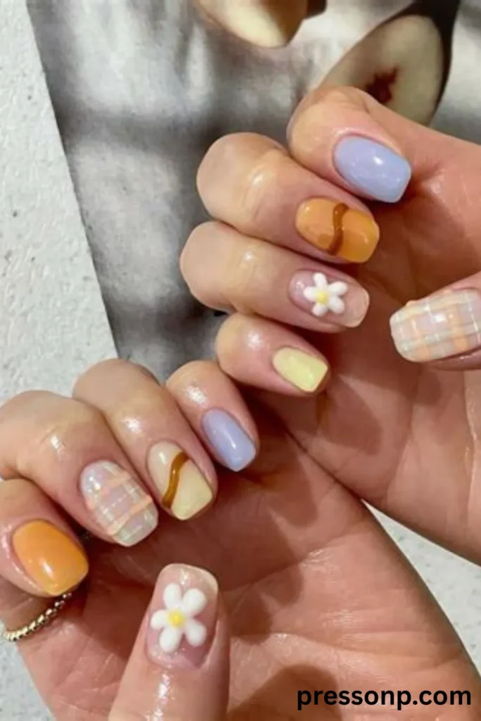 A close-up of two hands showing a playful, mismatched spring manicure on short squoval nails, featuring an assortment of solid pastel colors, 3D white daisy charms, a wavy brown line accent, and a soft pastel plaid pattern.