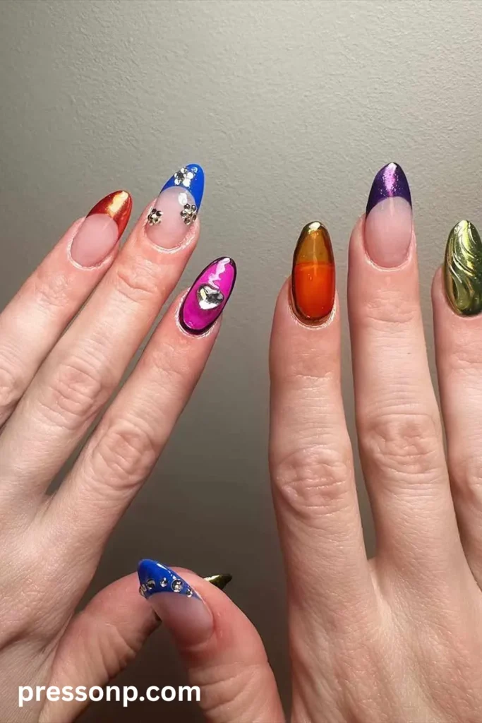 Close-up of almond-shaped nails showcasing an eclectic mix-and-match manicure with multi-colored metallic French tips, 3D chrome textures, rhinestone floral charms, and a prominent crystal heart accent.